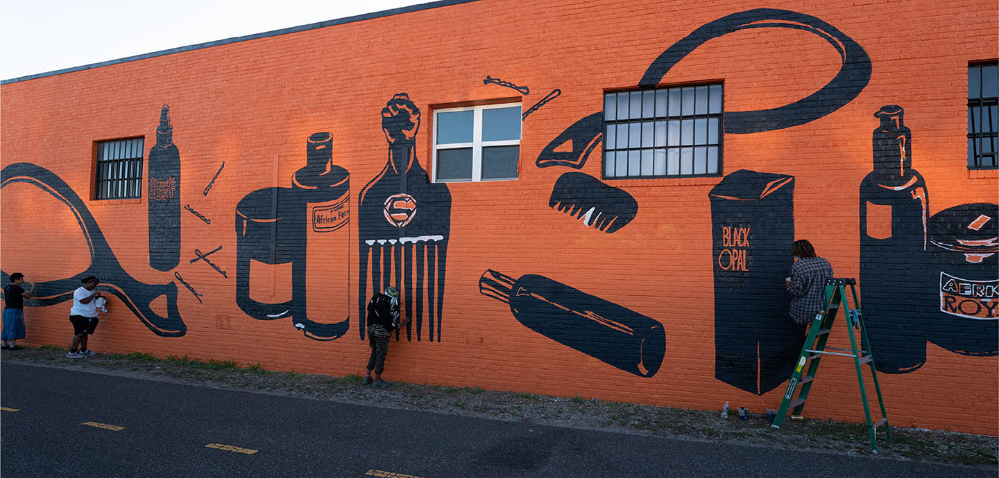 an orange-brown building with murals of hand mirrors and hair supplies painted in black. Four people stand in various places along the building, working on the mural. 