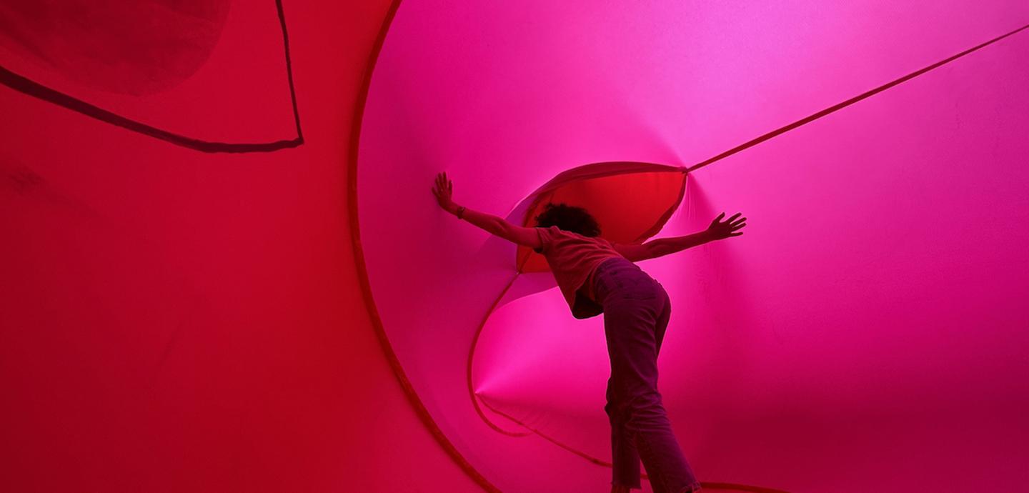 A large pink and red textile installation, with a person standing in the middle, their arms outstretched, as they lean forward.