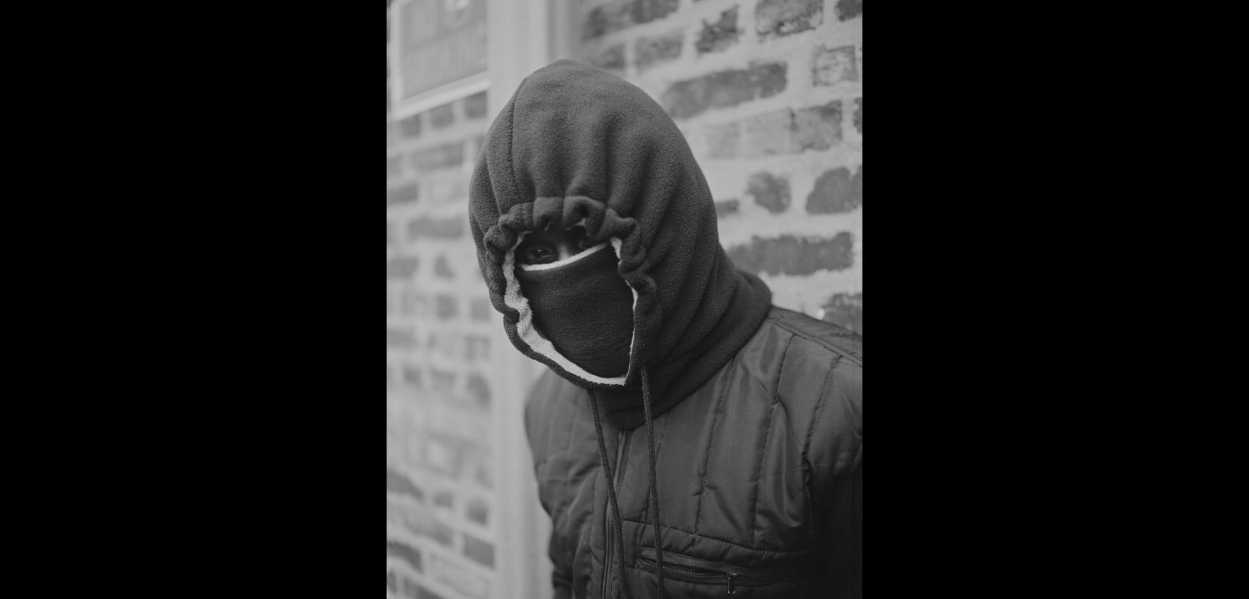 Black and white photo of a person wearing a dark colored hood and jacket with a thick mask covering face, leaning against brick wall.