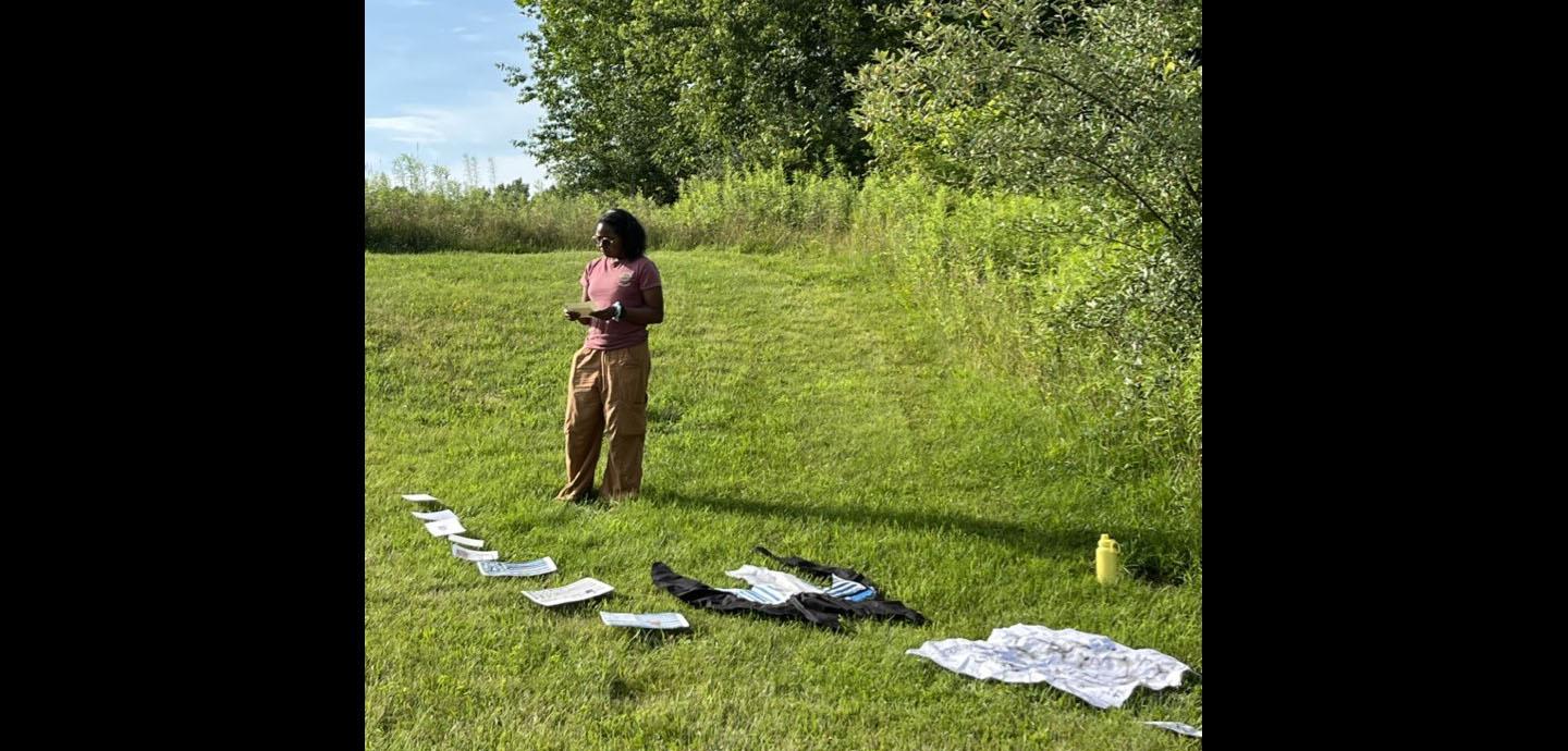Woman outside surrounded by green grass and trees with paper projects laid out on the ground in front of her.