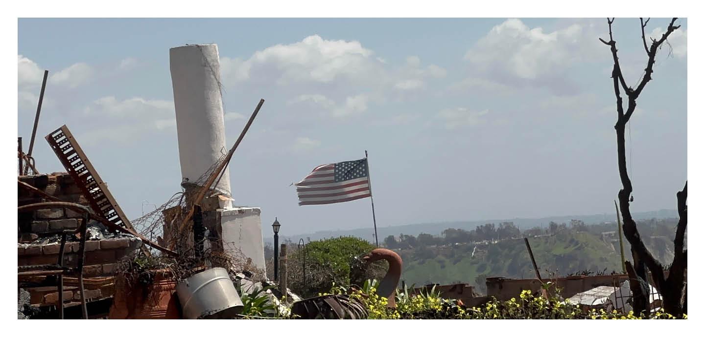 The broken remains of a house and an American flag, overlooking a hazy, charred landscape.