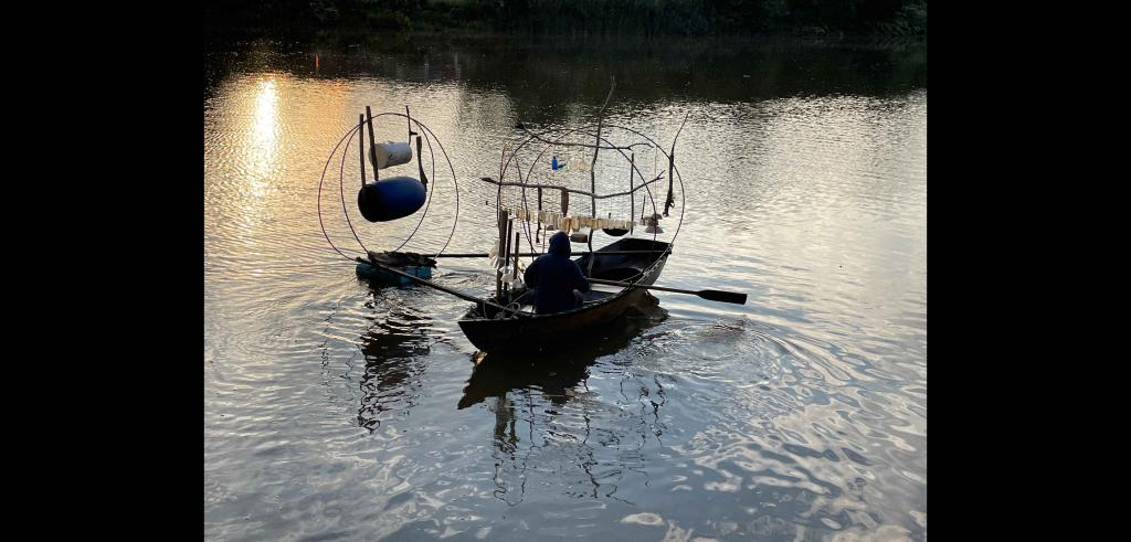 Artist sitting in a small rowboat with abstract sculptures attached, rowing in a body of water in dim lighting.