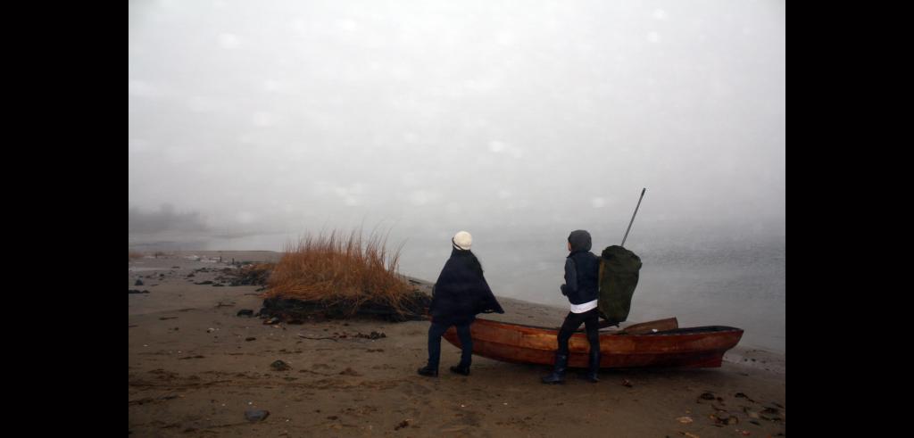Two people with their backs to the viewer, standing next to a small row boat on a shore under dark grey skies. The person on the left is wearing a cape and the person on the right is wearing a hooded rain jacket and large backpack with an antena sticking out of it. 