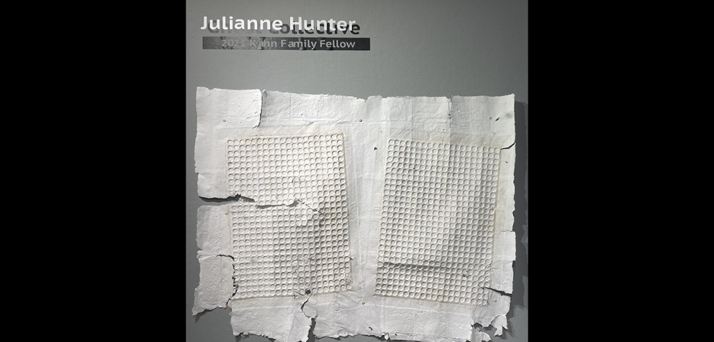 Against a light grey wall hangs a piece of handmade paper with irregular edges and embossed patterns of repeated circles. 