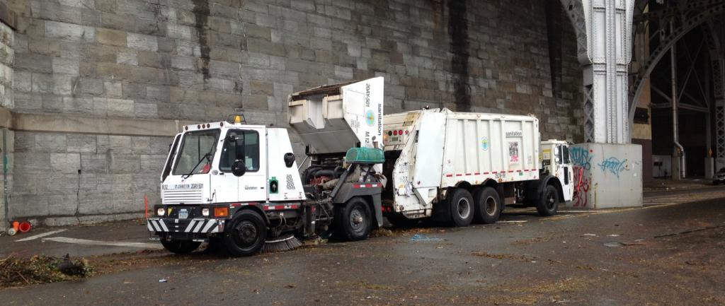 A white garbage truck in front of a concrete wall under an overpass.