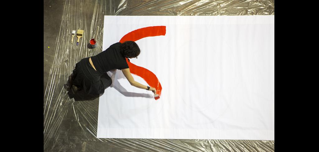 Person wearing black clothes, kneeling on the ground, painting a red curved line on a large white canvas on the floor next to a paint can and paint brushes.
