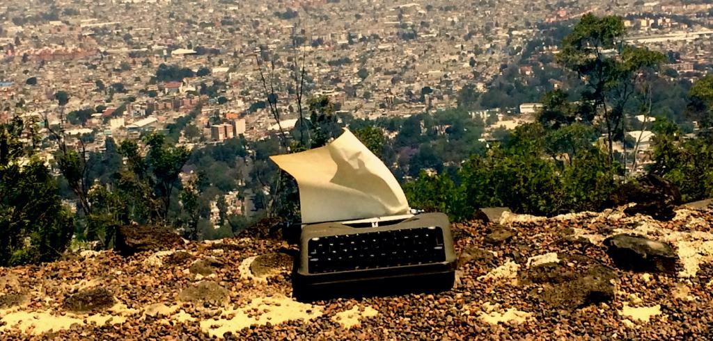 Typewriter on the ground with a blank piece of paper hanging from the top overlooking a city with trees in the distance.