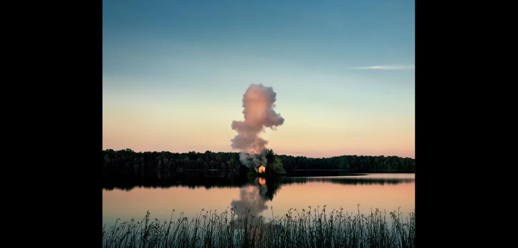 A smoking house surrounded by a lake at sunset with pastel colors of blue, yellow and peach reflected in the water.