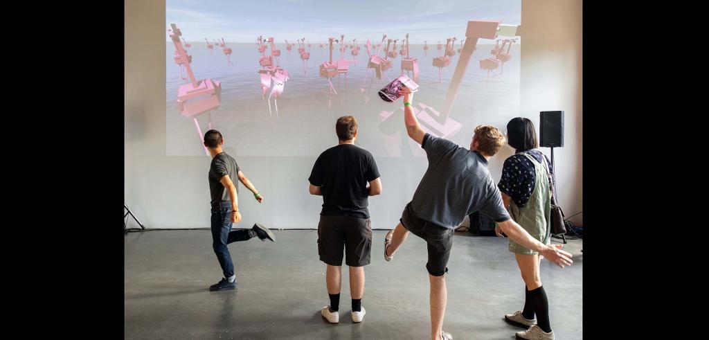 Four people looking at a projection of pink abstract flamingos with two people standing on one leg in front.