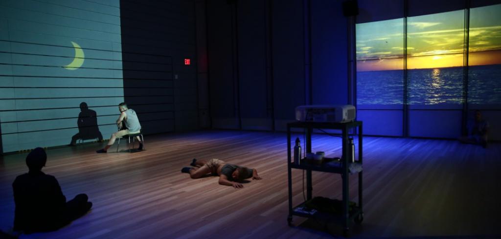 Two projections in a dark space, one of a crescent moon, the other of a sunset on water, with one man sitting in a chair, one person on the ground, one person in shadow.