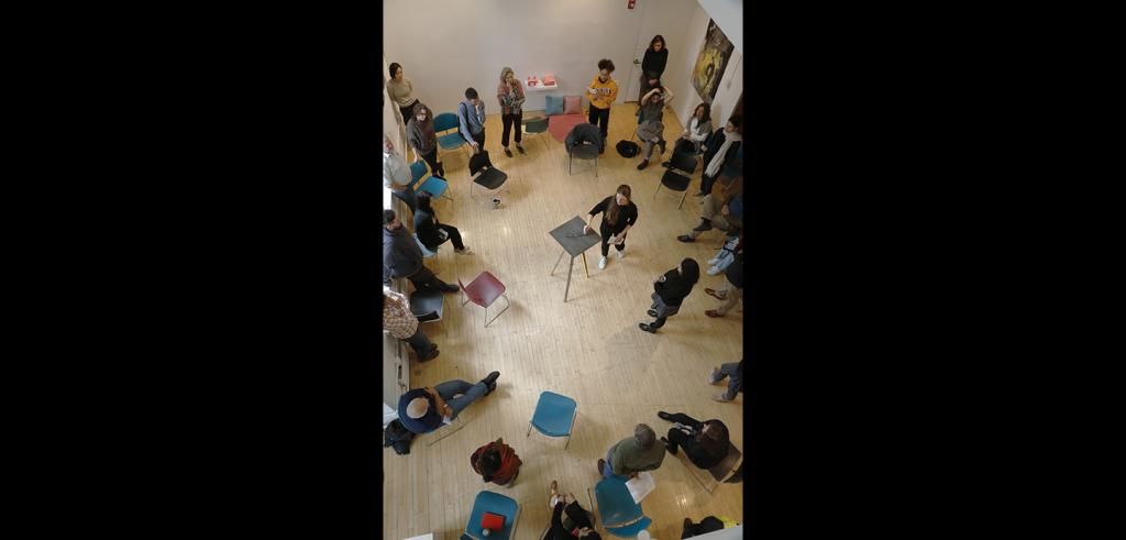 Group of people in a loose circle looking at a person presenting artwork.