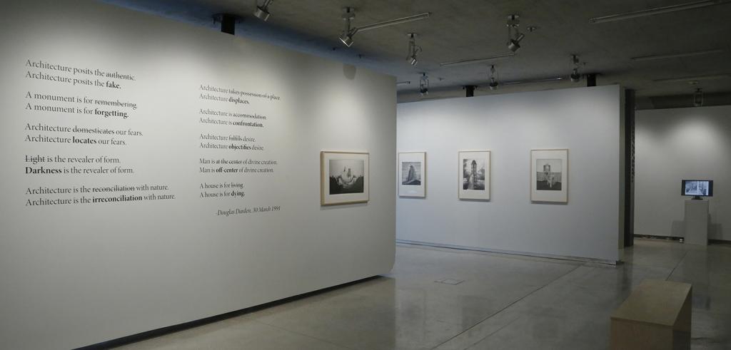 View of an art gallery with framed images and text on white walls and monitor on a pedestal.