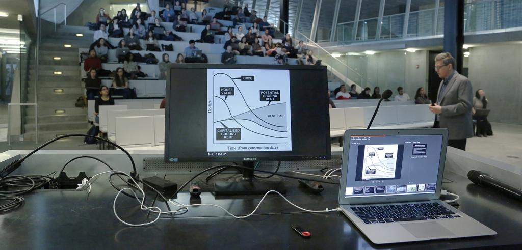 Two computer screens on a podium, with people sitting in an auditorium and man walking on right.