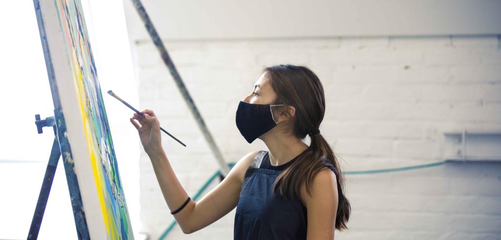 Woman with long brown hair pulled back wearing a black face mask, painting on an easel.