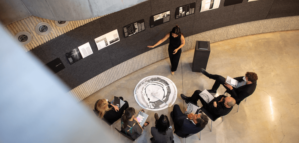 People sitting in chairs observing a person showing artwork on a wall.