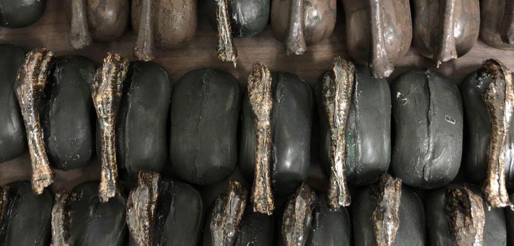 Round rectangular objects in dark brown and dark grey arranged in three rows with skinny bronze pieces placed randomly on top.