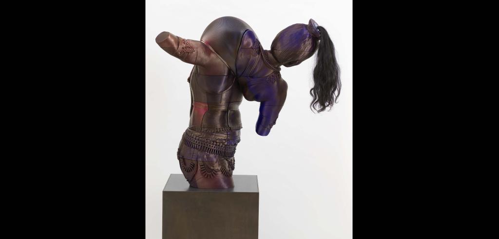 Rear view of previous bust of a figure with long dark hair leaning to the left wearing an intricate outfit and carrying a large dome in front of it on a steel pedestal.