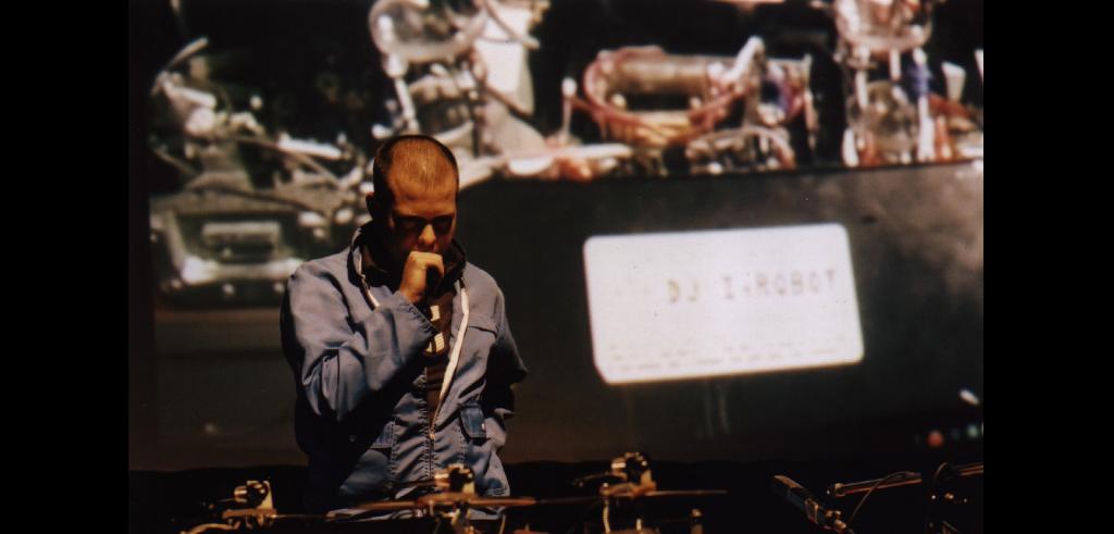 Person with a shaved head holding a microphone wearing a long sleeve dark shirt on a stage.