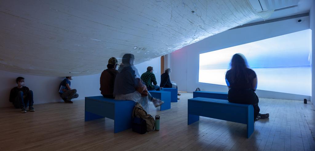 People sitting in a dark room on white benches looking at a body of water projection sitting underneath a long piece of fabric.