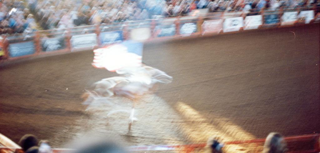 Blurred video still of a person riding a bull in a large pen.