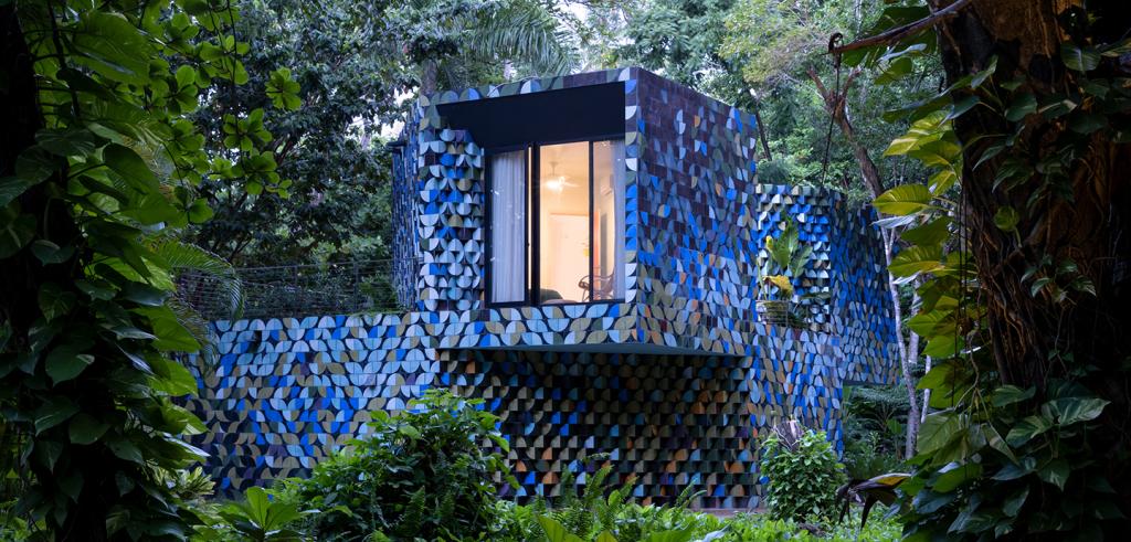 A house composed of stacked and blue multi-colored concrete masonry blocks, foregrounded by a vibrant green jungle landscape. 