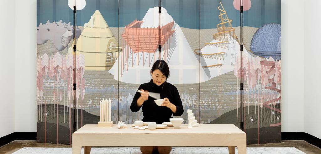 A woman behind a table holding small ceramic pieces with a mural of mountains behind her.