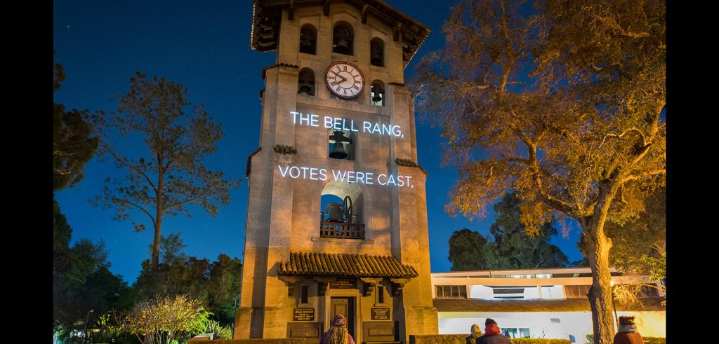 A tower with a lit up clock near the top with the words The Bell Rang, Votes Were Cast over the tower, image taken near dusk.