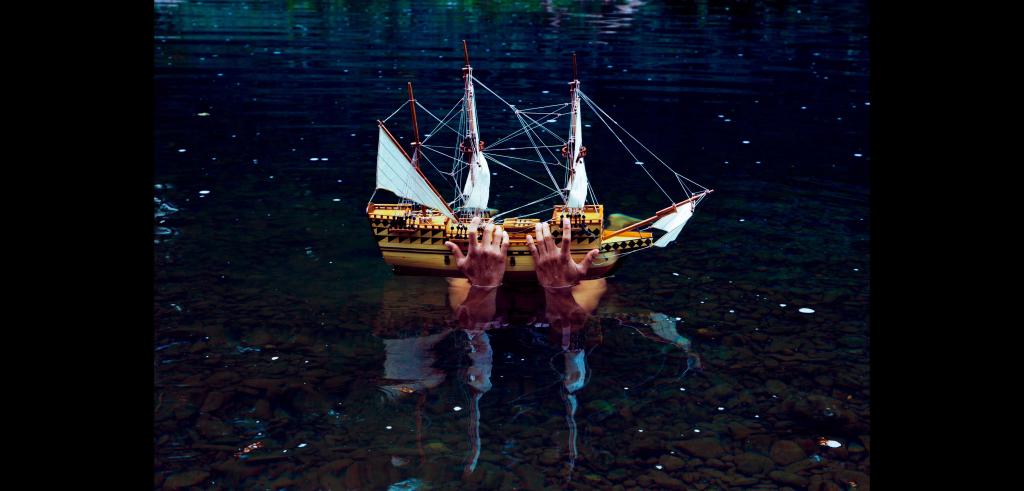 Two hands holding a small model boat with sails (about one and a half feet long) above water in the nighttime.
