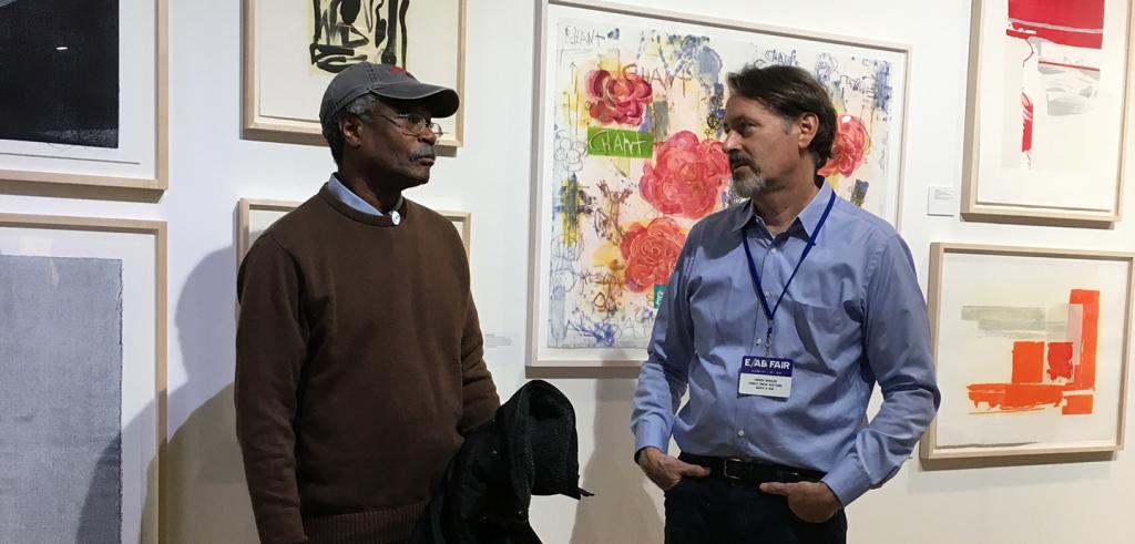 Two men converse in front of a wall of framed artwork
