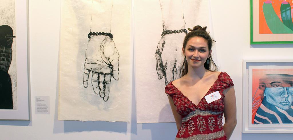 Woman with long hair stands beside two drawings of hands hung on a gallery wall