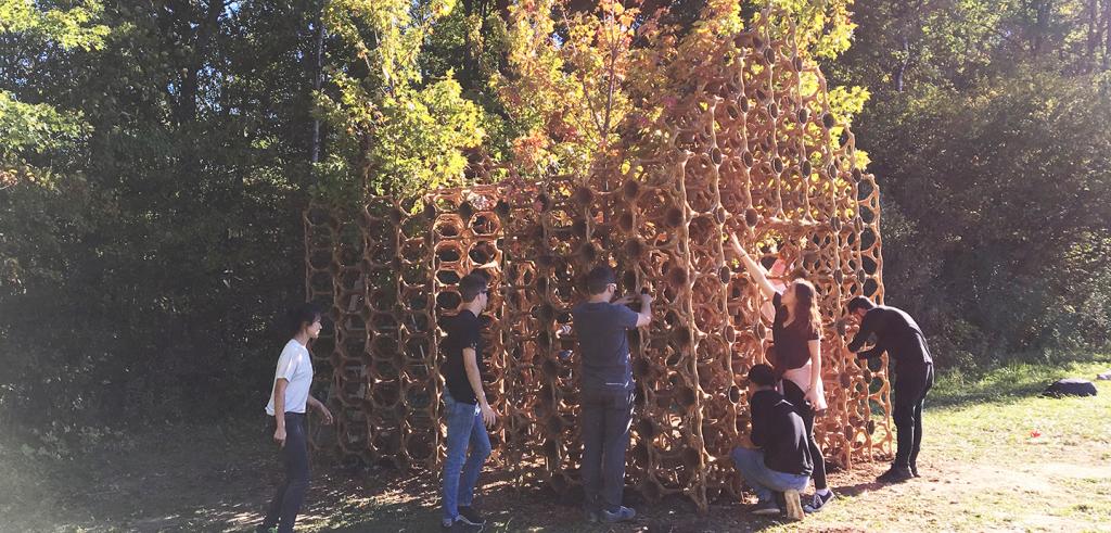 Wooden structure made of circular components set in a field with trees behind it