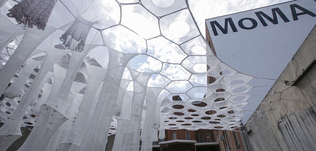 woven white structures creating a canopy in a courtyard with a building with MoMA painted on it in the background