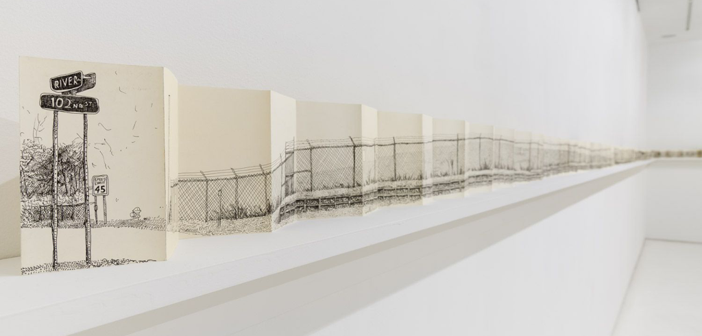Photo of a gallery in which a single piece of long paper is spread along a ledge on the white walls. Drawn on the paper is a chainlink fence and street signs. 
