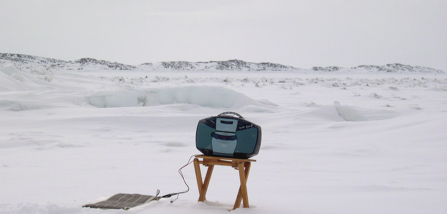 A black and blue boombox sits atop a wooden folding stool against the backdrop of a snowy tundra. 