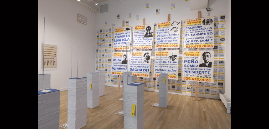 Photo of an art installation consisting of numerous posters full of blue, yellow, and red Spanish text and various black printed images. Stacks of paper multiple feet high lay on the light wood floor in front of the posters hung on a white gallery wall. 