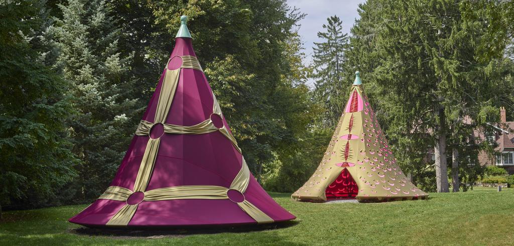Two fabric tents, one pink and gold the other red and gold on grass surrounded by trees and a house in background.