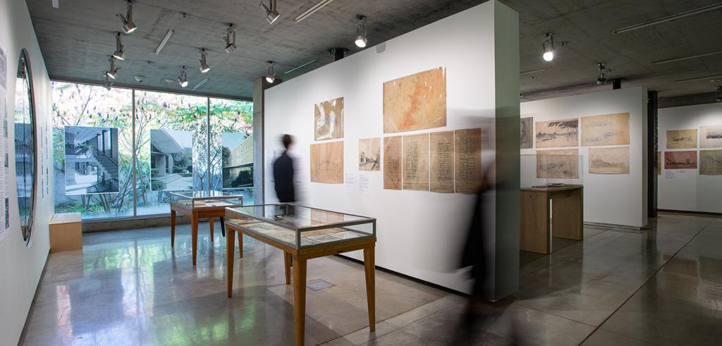 Art gallery with pieces displayed on walls, windows and in glass cases. A blurry person is looking at the wall hangings.  