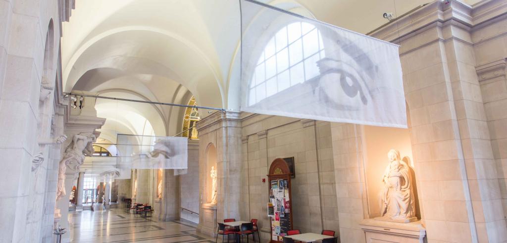 Inside of a light gray building lit with a light yellow hue with glass projections featuring a human eye throughout the hallway.