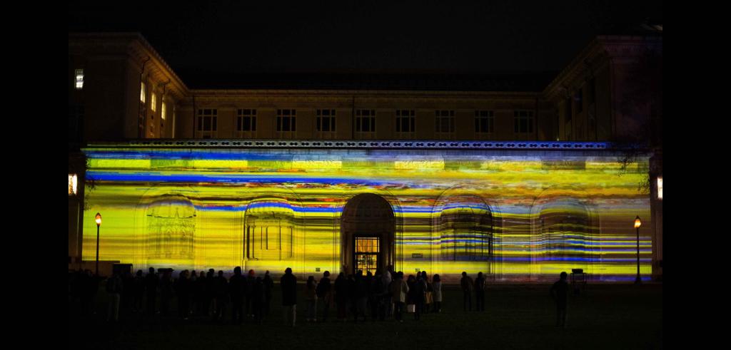 Illuminated building with bright stripes of yellow, blue, and some orange with a group of onlookers in front at night.