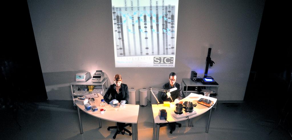 Two people sitting at semicircular desks in front of a projected image of DNA.