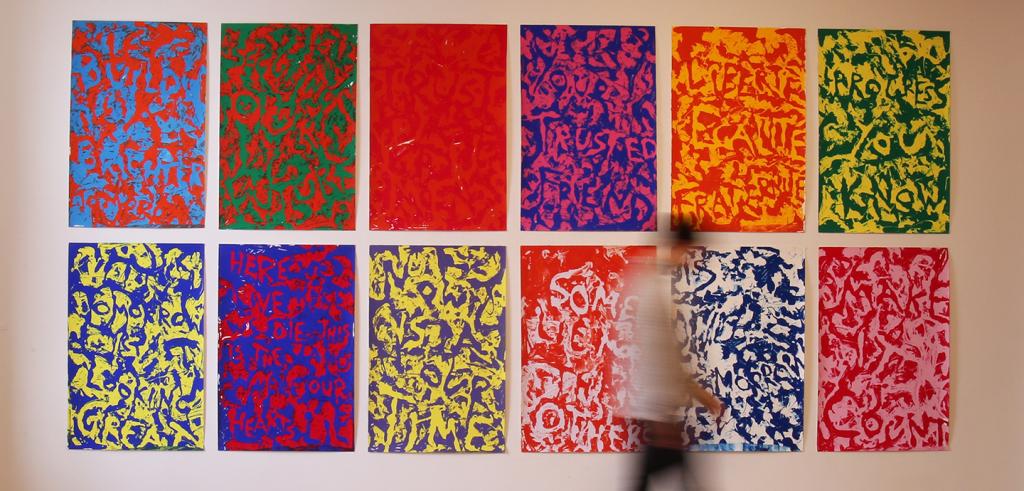 A series of 12 colorful posters mounted on a wall with a person moving in front of them