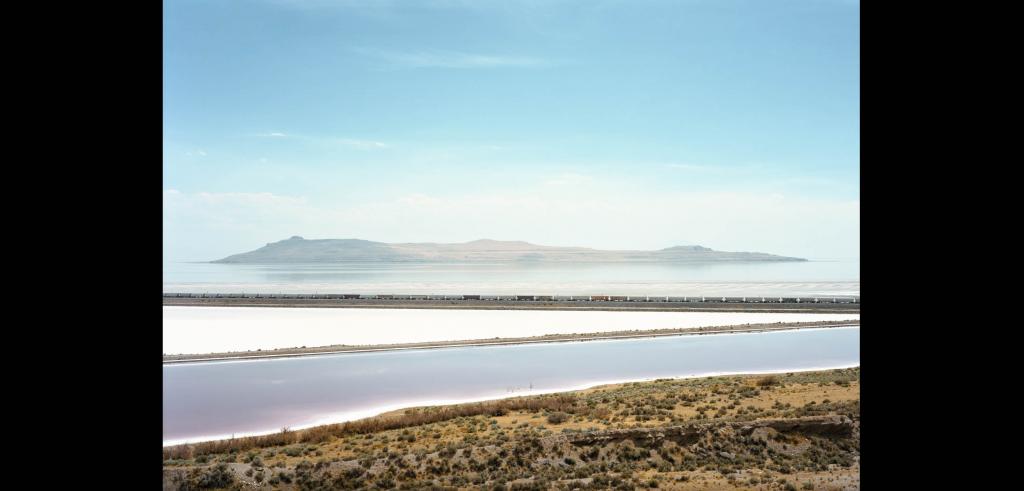 A lake with part of a desert on the bottom right corner with a large light blue sky above it with a train across the middle.