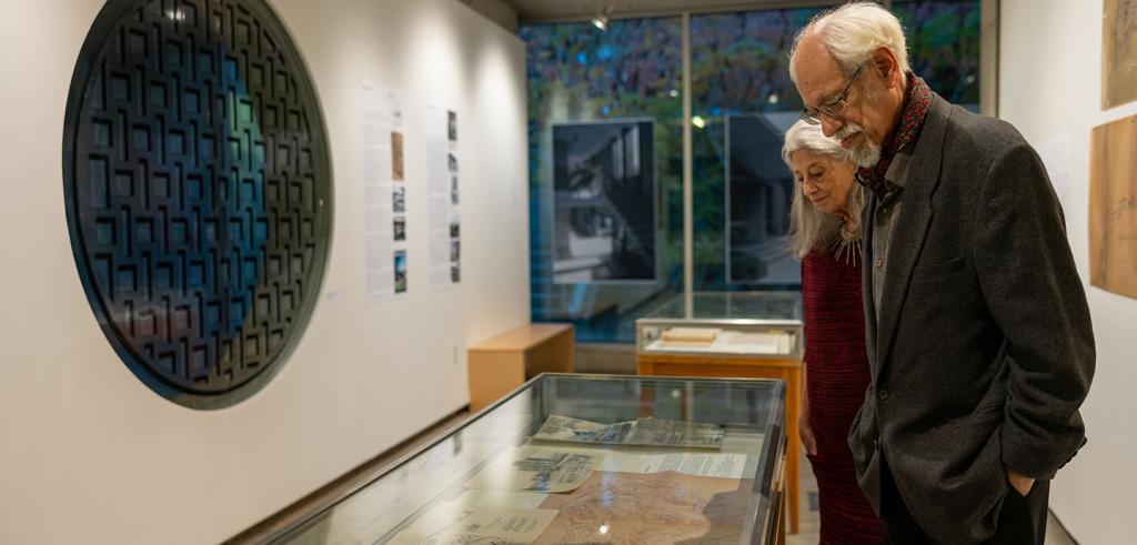 Man with jacket, colorful scarf and glasses along side a woman wearing a red dress and silver necklace looking at drawings in enclosed glass casing in an art gallery   