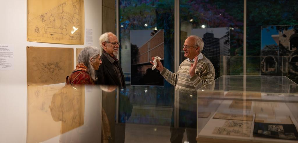 Three people in conversation inside an art gallery. Wall hangings, window hangings and enclosed glass cases are present. 
