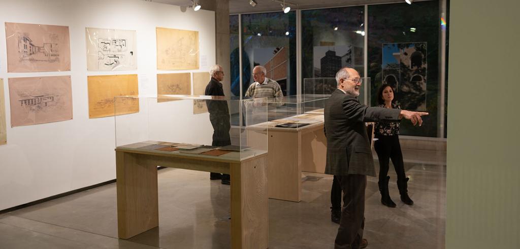 A group of four inside an art gallery with wall hangings and peinces in enclosed cases. Man in sut and glasses pointing at piece on the wall next to a woman in black pants and floral top. In the back two mean are in conversation. 