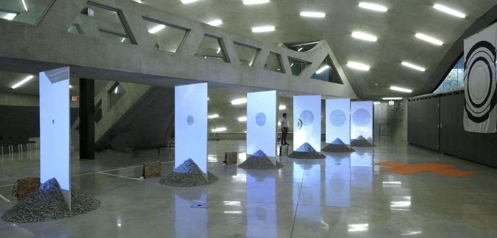 Six illuminated vertical boards with low gravel piles at their bases, in a gallery space
