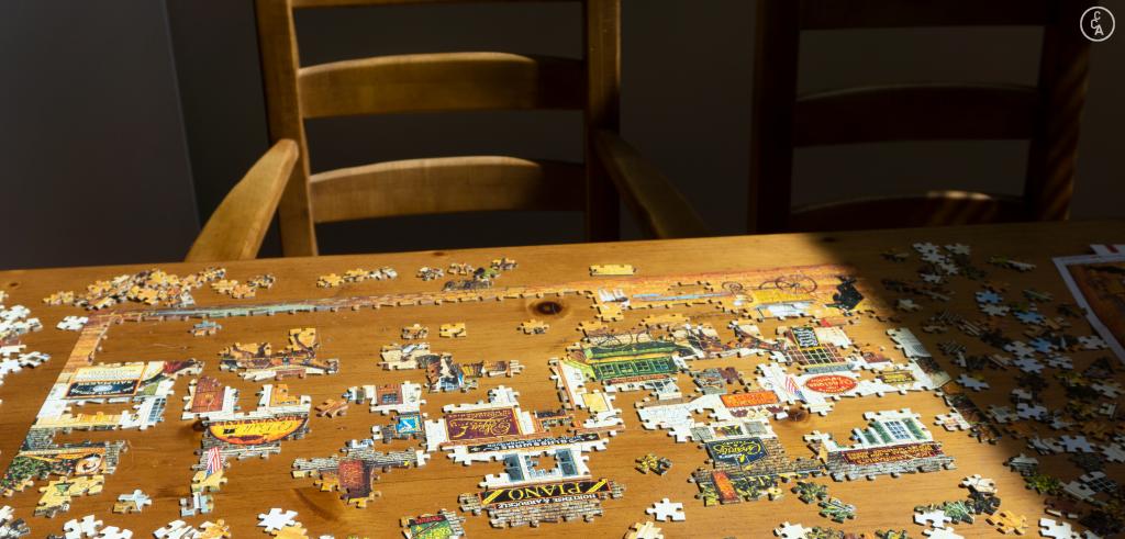 Puzzle partially put together on a wooden table with a wooden chair in a shadow.