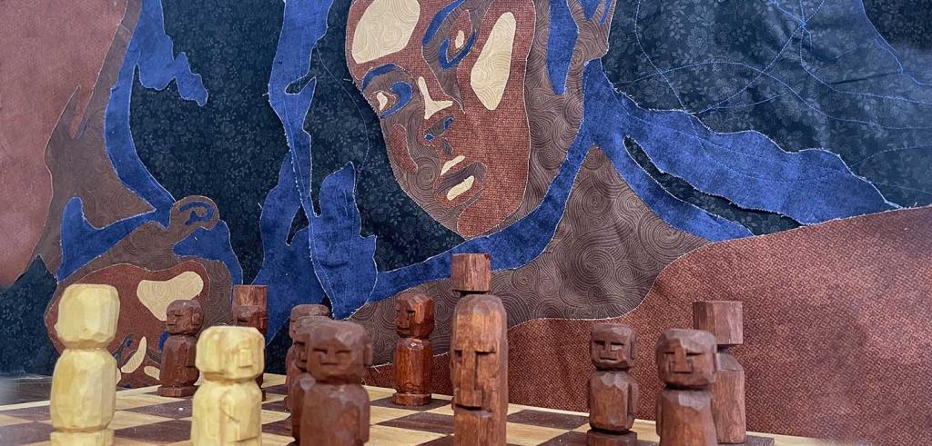 Abstract painting of a woman with dark blue hued hair with brown pigments behind a hand carved wooden chess set.