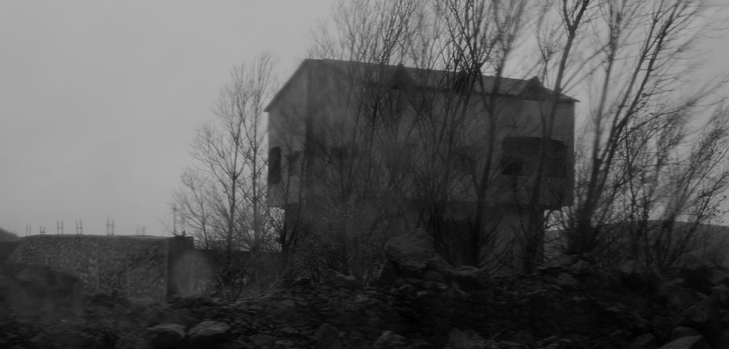 Black and grey photo of a simple house on a tall foundation, surrounded by boulders and rock rubble, with a foreground of barren trees. 