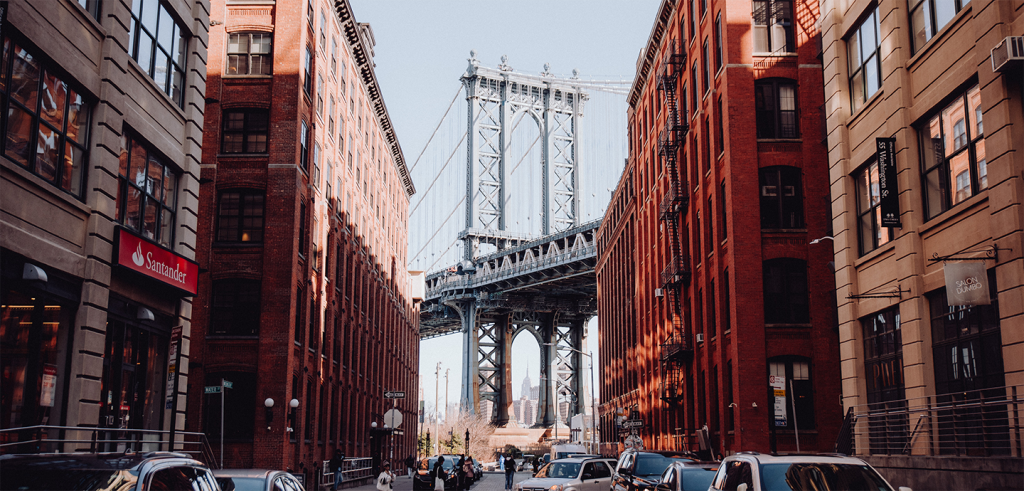 A city street view with buildings, cars, and people on the street with a bridge in the background.